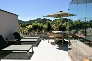 A rooftop terrace with outdoor furniture, a glass wall, and a view of green hills. - Casa Marina Piccola (São Sebastião)