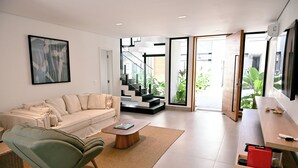 Una sala de estar moderna con un sofá, un sillón verde, una mesa de centro y una escalera que conduce a un balcón con plantas.