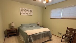 Een slaapkamer met een bed, twee nachtkastjes, een stoel, een raam met zonwering en een plafondventilator.