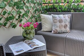 A wicker sofa with a cushion, a glass vase with pink flowers, and a magazine on a small table. - SAN GIOVANNI Appia Park - Stylish apartment with balcony - Metro A - 10 min City Center (Rome)
