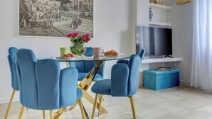 A dining area with a glass table, blue chairs, a painting, and a television on a white stand. - SAN GIOVANNI Appia Park - Stylish apartment with balcony - Metro A - 10 min City Center (Rome)