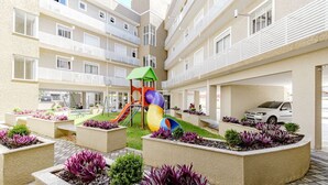A multi-story residential building with balconies, a children's play area with slides, and landscaped planters with purple foliage. - Apto com 2 Vagas e Churrasqueira Ingleses (Florianópolis)