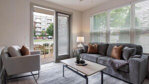 A modern living room with a grey sofa, a small armchair, and a coffee table. There is a window with blinds, a lamp, and a view of a residential area outside. - Landing Apartments Near Autumnwood (Charlotte)