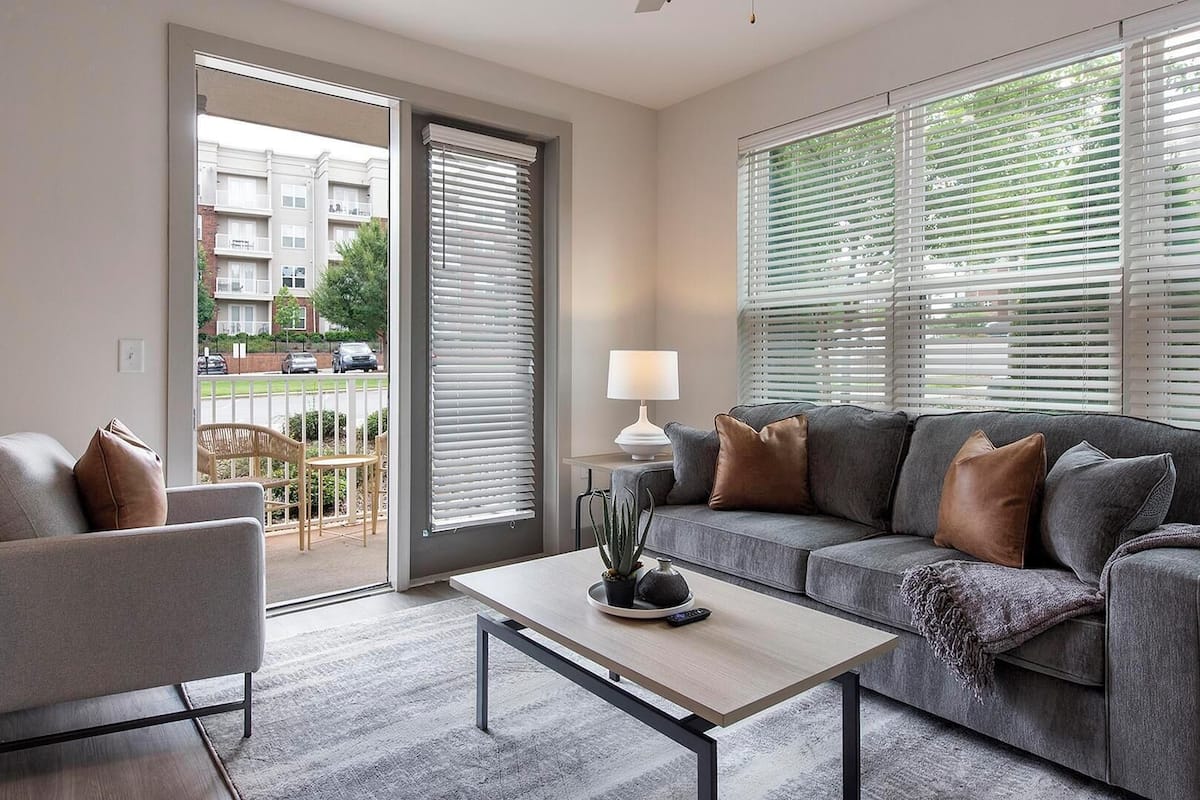 A modern living room with a grey sofa, a small armchair, and a coffee table. There is a window with blinds, a lamp, and a view of a residential area outside.