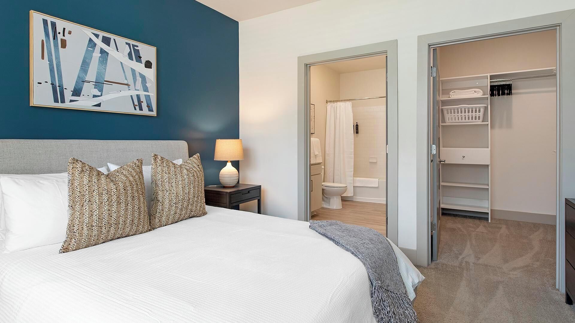 A hotel room with a large bed, a nightstand with a lamp, a bathroom visible through an open door, and a walk-in closet.