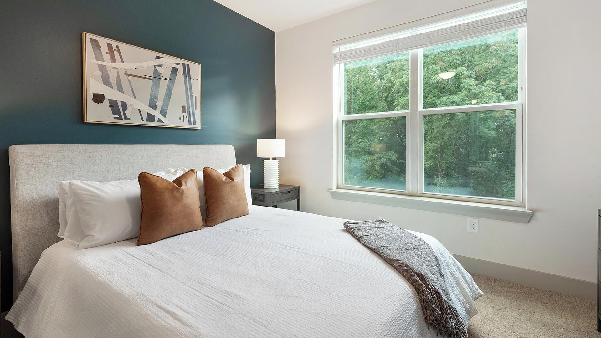 Een slaapkamer met een groot bed, een nachtkastje, een lamp en een raam met uitzicht op bomen.