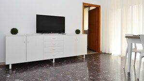 Una stanza moderna con un armadietto bianco, una TV a schermo piatto, una piccola pianta, una porta in legno e una sedia bianca.