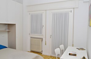 Quarto com cama, escrivaninha com cadeiras, radiador e guarda-roupa com cortinas.