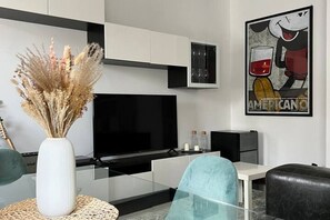Un salon moderne avec un téléviseur à écran plat, un vase contenant des fleurs séchées, une table basse en verre et un tableau encadré accroché au mur.