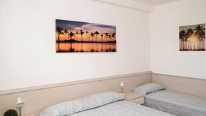 A hotel room with two beds, a nightstand, a lamp, and two wall-mounted paintings of palm trees at sunset. - Cattel A05 with Private Terrace and Air Conditioning (Lido di Jesolo)