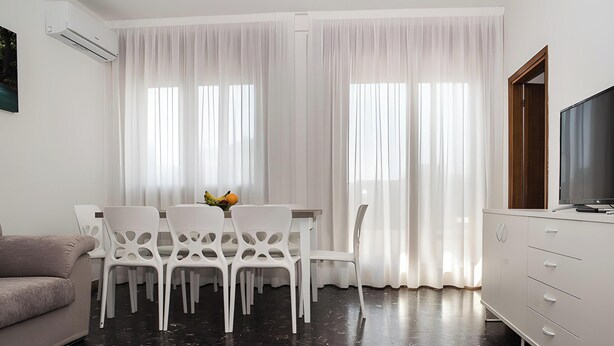 A modern living room with a sofa, dining table with chairs, a TV, and a large window with sheer curtains. - Cattel A05 with Private Terrace and Air Conditioning (Lido di Jesolo)