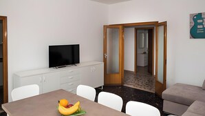 A room with a dining table, chairs, a TV on a cabinet, a sofa, and a door leading to another room. - Cattel A05 with Private Terrace and Air Conditioning (Lido di Jesolo)