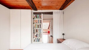 A bedroom with a wooden ceiling, a white bed, a bookshelf, and a red chair in the background. - Panoramic apartment with a view of the Po River (Torino)