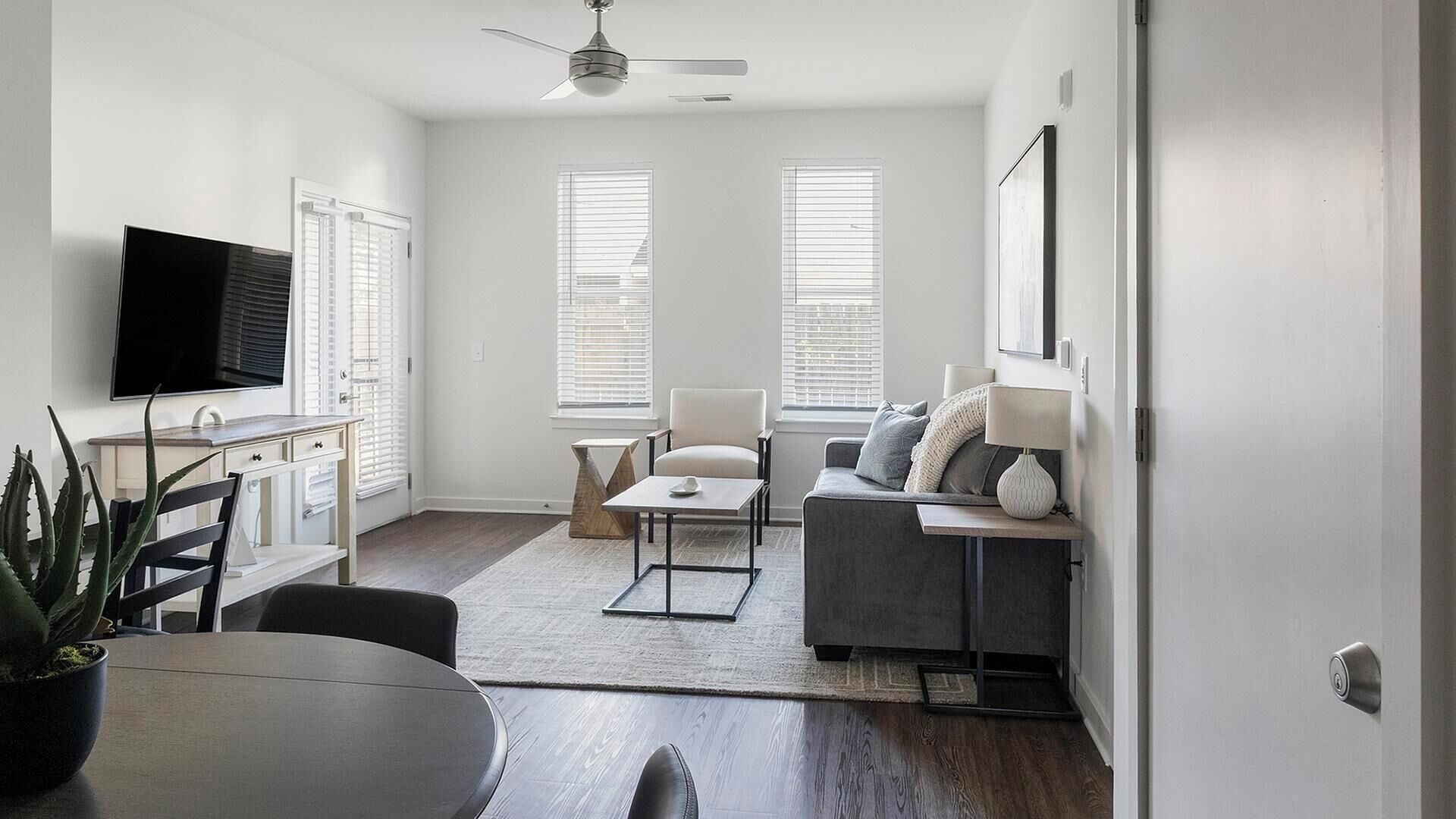 Un salon moderne avec un canapé gris, une table basse et un téléviseur fixé au mur. Il y a deux fenêtres équipées de stores, un ventilateur de plafond et une plante en pot.