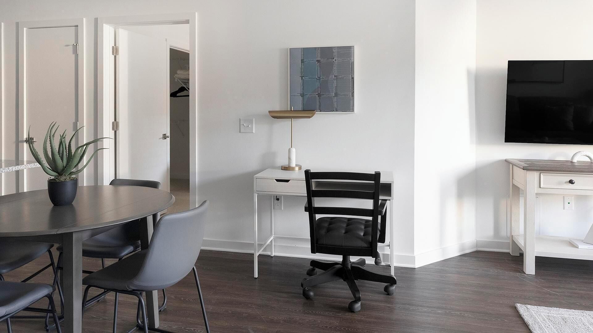 A modern living room with a dining table, chairs, a black office chair, a small desk with a lamp, a TV, and a potted plant.