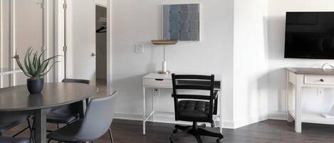 A modern living room with a dining table, chairs, a black office chair, a small desk with a lamp, a TV, and a potted plant.