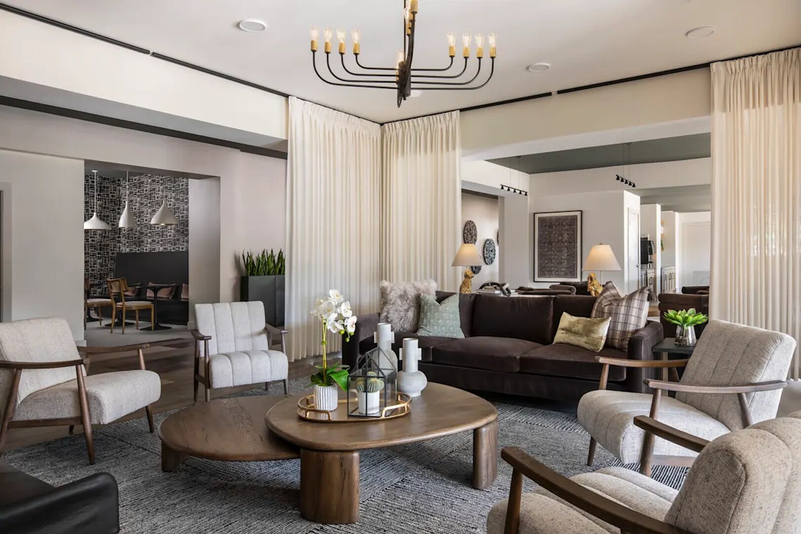 Uma sala de estar moderna com uma mesa de centro redonda, sofás e cadeiras. É possível ver um candelabro, uma área de refeições e uma cozinha ao fundo.