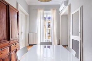 Ein Zimmer mit einem Holzkabinett, einem weißen Tisch und Blick durch ein mit Vorhängen versehenes Fenster.