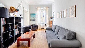 Ett vardagsrum med en grå soffa, ett träbord, en svart bokhylla och ett fönster med gardiner.