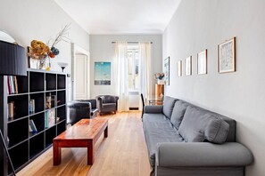 Ein Wohnzimmer mit grauem Sofa, einem Holztisch, einem schwarzen Bücherregal und einem Fenster mit Vorhängen.