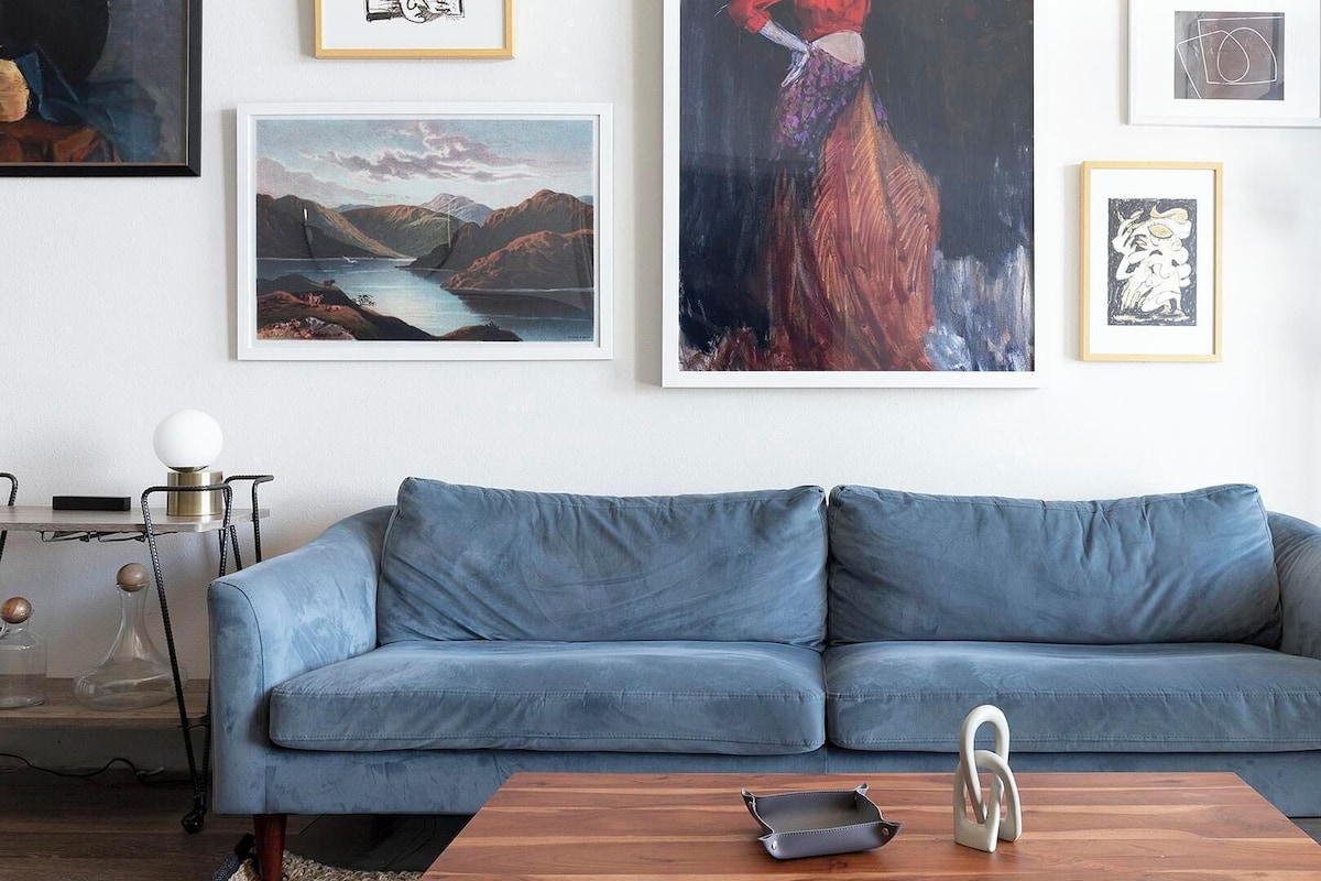 A modern living room with a blue sofa, a wooden coffee table, and various framed artworks on the wall.