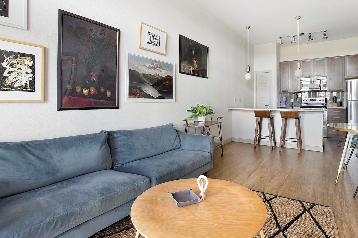 A modern living room with a grey sofa, a round wooden coffee table, and a kitchen area with stainless steel appliances and a bar stool.