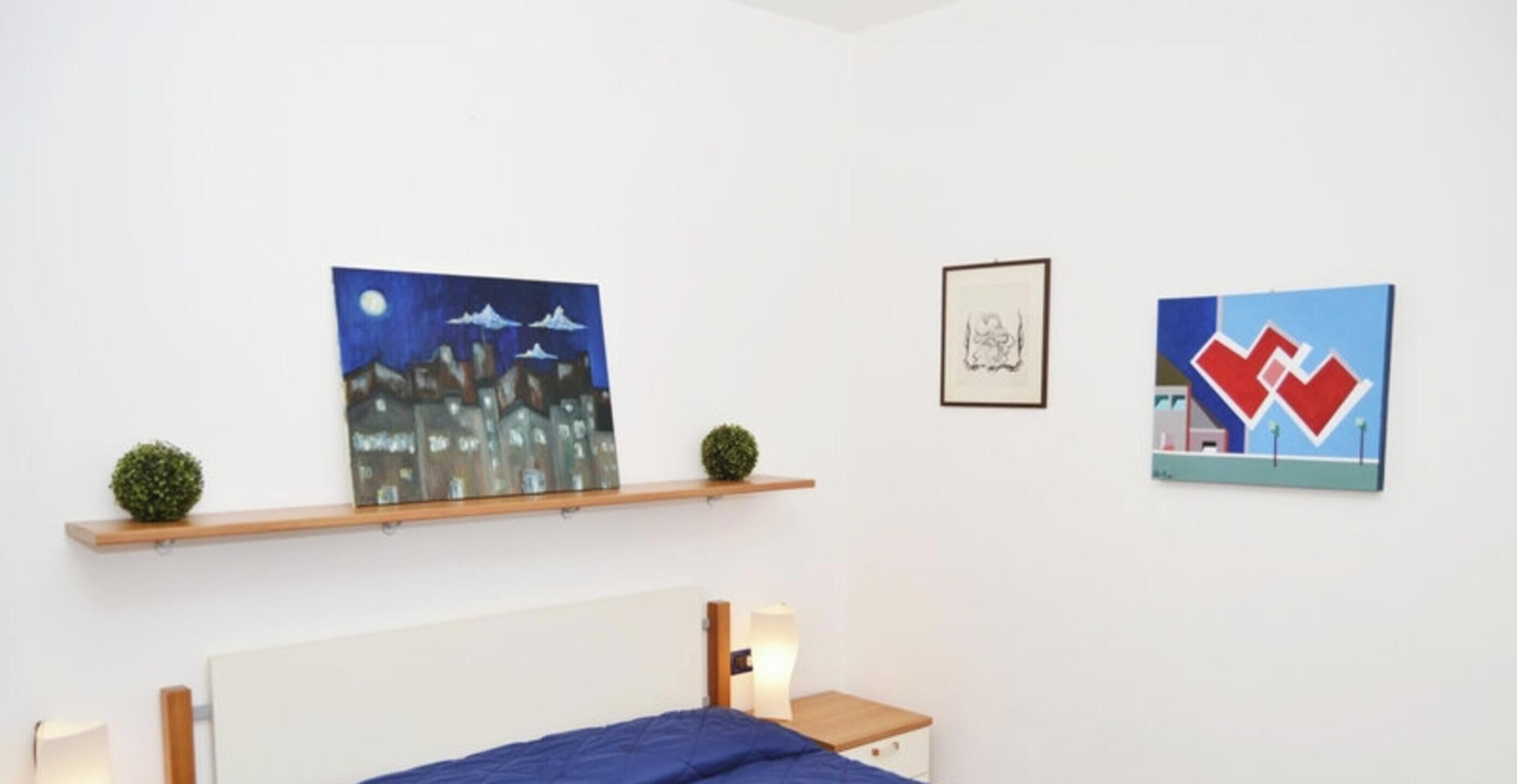 A bedroom with a bed, a nightstand, a shelf with artwork, and a framed picture on the wall.