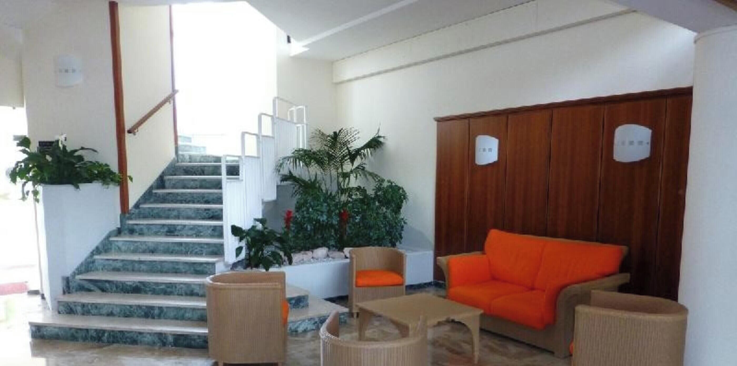Un hall de hotel con una escalera, muebles de mimbre, un aparador de madera y plantas en macetas.