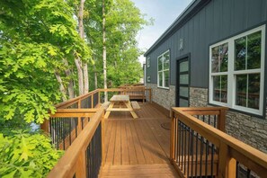 A wooden deck with a picnic table, surrounded by trees and a building with stone walls and large windows. - Greystone (Anderson)