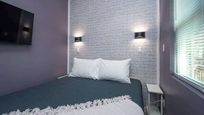 Ein Schlafzimmer mit einem Ziegelstein-Muster-Kopfende, zwei Wandleuchten, einem Bett mit weißen Kissen, einer weißen Tagesdecke und einem Fenster mit Jalousien.