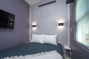 Une chambre à coucher avec une tête de lit ornée d’un motif en briques, deux lampes fixées au mur, un lit avec des oreillers blancs, un plaid blanc et une fenêtre avec des stores.
