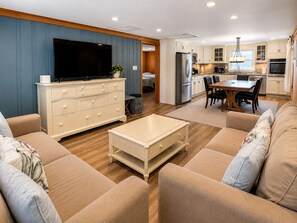 A modern living room with a flat-screen TV, beige sofas, a coffee table, and a kitchen in the background. - Walk to Beach! Cozy 2BR w/ Huge Private Deck + Parking (Ocean City)
