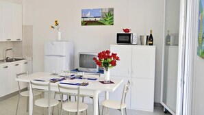 Eine kompakte Küche mit weißen Schränken, einem Esstisch mit Stühlen, einer Mikrowelle, einem kleinen Kühlschrank und einer Vase mit roten Blumen.