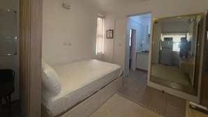 A bedroom with a bed, a mirror, a small window, and a door leading to a kitchen. - Cozy 1-bedroom apartment in peaceful Lagos with AC, gym and pool access (Lagos)