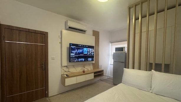 A hotel room with a bed, a TV mounted on the wall, a wardrobe, and a refrigerator. - Cozy 1-bedroom apartment in peaceful Lagos with AC, gym and pool access (Lagos)