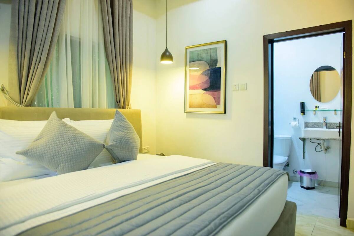 Habitación de hotel con cama, almohadas, un cuadro, una lámpara de noche y un baño visible a través de una puerta abierta.