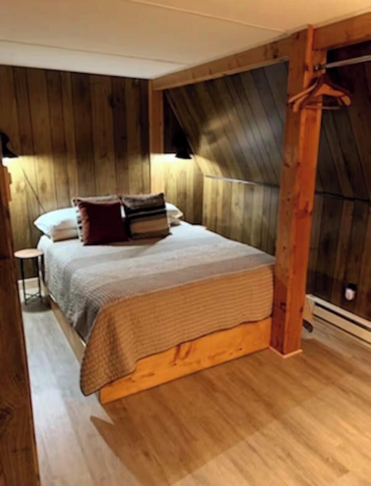 Une chambre avec un plafond en pente en bois, un lit recouvert d’une couverture grise et un plancher en bois.