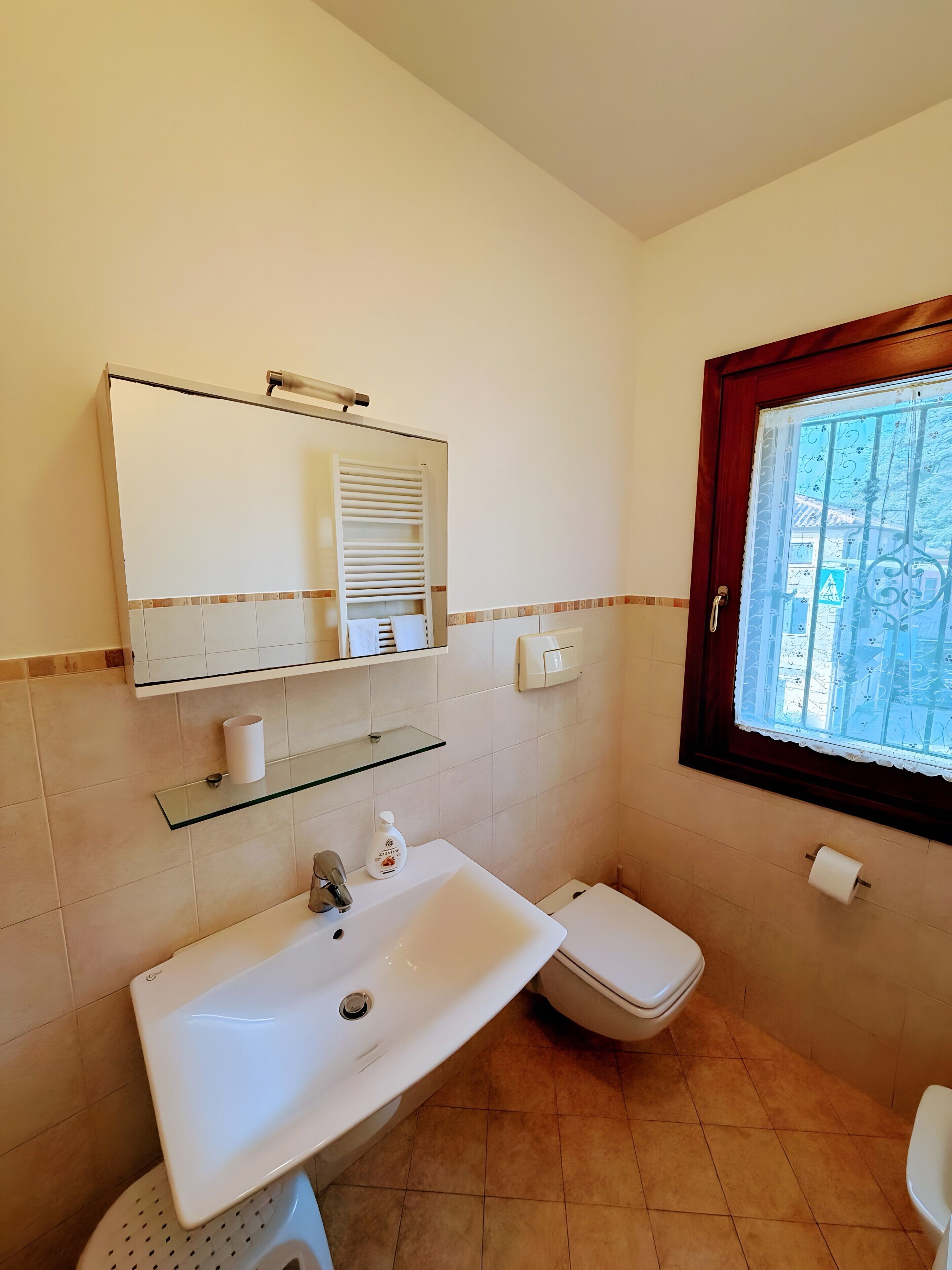 A bathroom with a white sink, a toilet, a glass shelf, a mirror, and a window with a decorative grille.