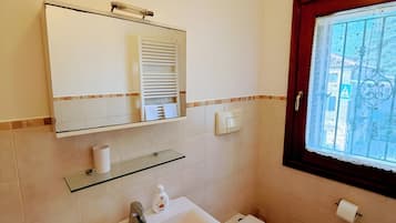 A bathroom with a white sink, a toilet, a glass shelf, a mirror, and a window with a decorative grille.