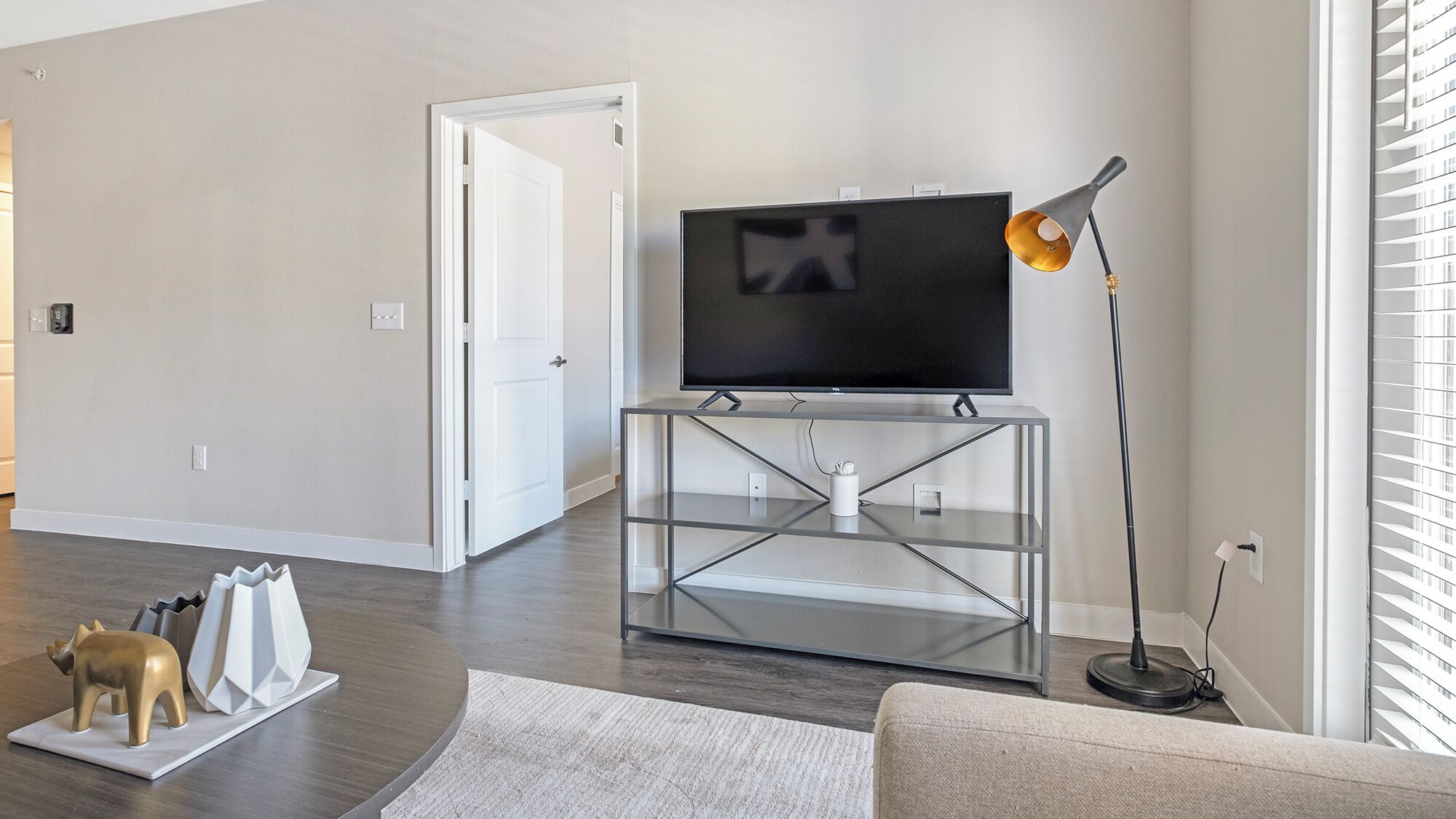 A modern living room with a flat-screen TV on a glass TV stand, a standing lamp, and a door leading to another room.