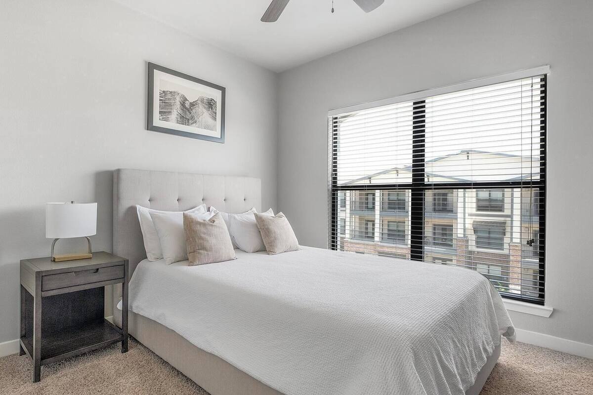 A bedroom with a bed, a nightstand, a lamp, a framed picture, and a window with blinds.