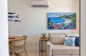 A room with a dining area, a sofa, a painting of a tropical beach, and an air conditioning unit. - Palmer | Jockey de Itaparica | Panoramic View (Vila Velha)
