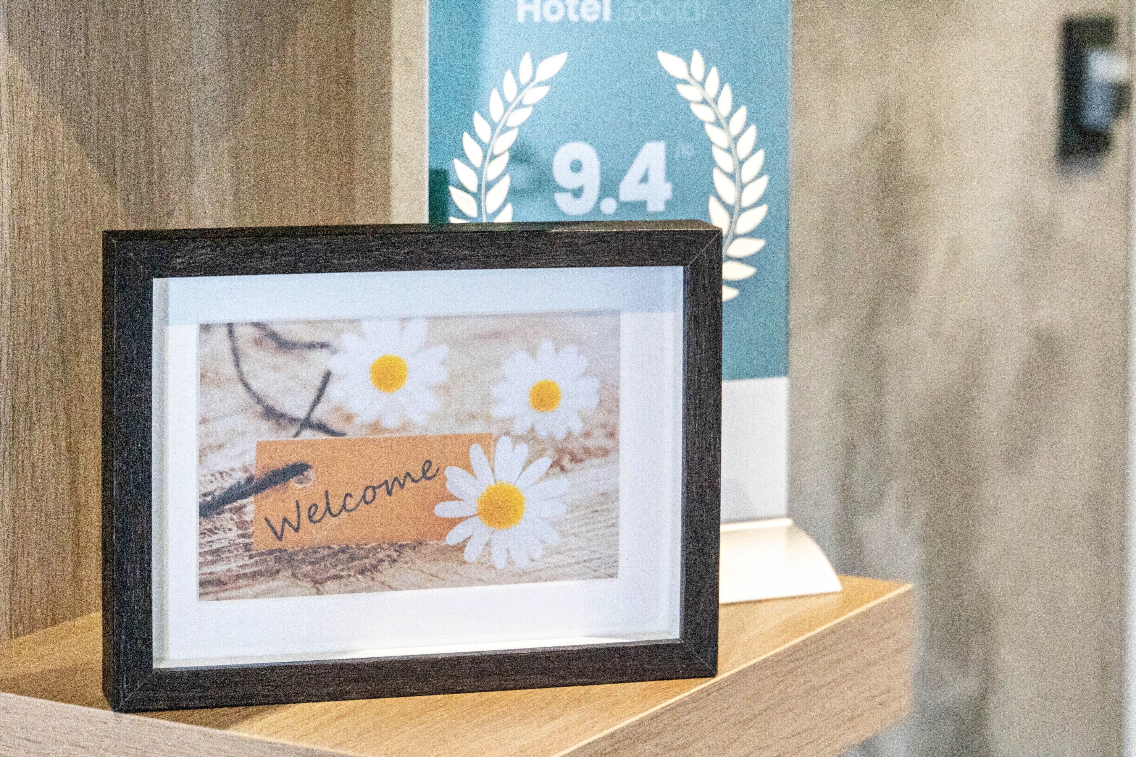 Ein gerahmtes Willkommen-Schild mit Margeriten und dem Schriftzug 'Welcome', ein Holzregal und ein Hotelschild im Hintergrund.