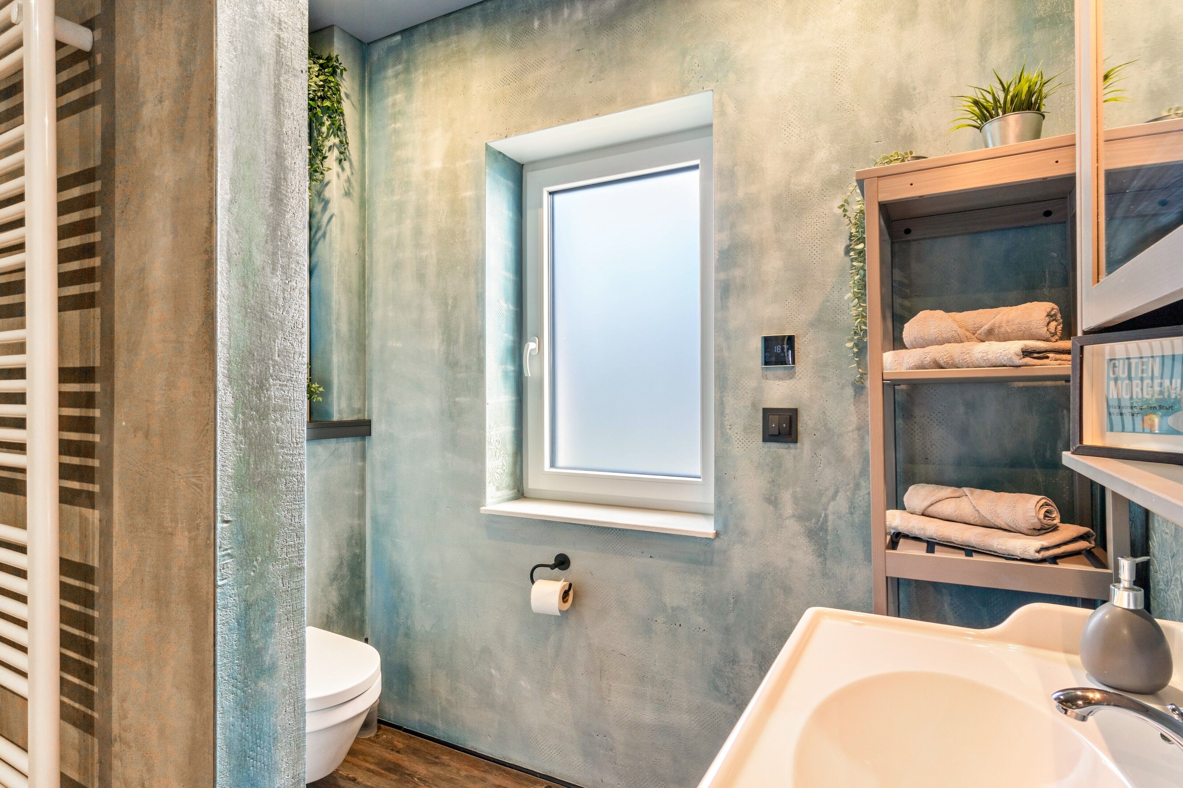A bathroom with a toilet, a sink, a towel rack, a window, and a shelf with towels.
