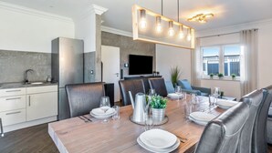 Un comedor moderno con una mesa de madera dispuesta para seis comensales, una cocina con armarios blancos y un gran ventanal con cortinas.