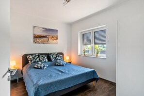 Un dormitorio con cama, mesita de noche, ventana con persianas y un cuadro colgado en la pared.