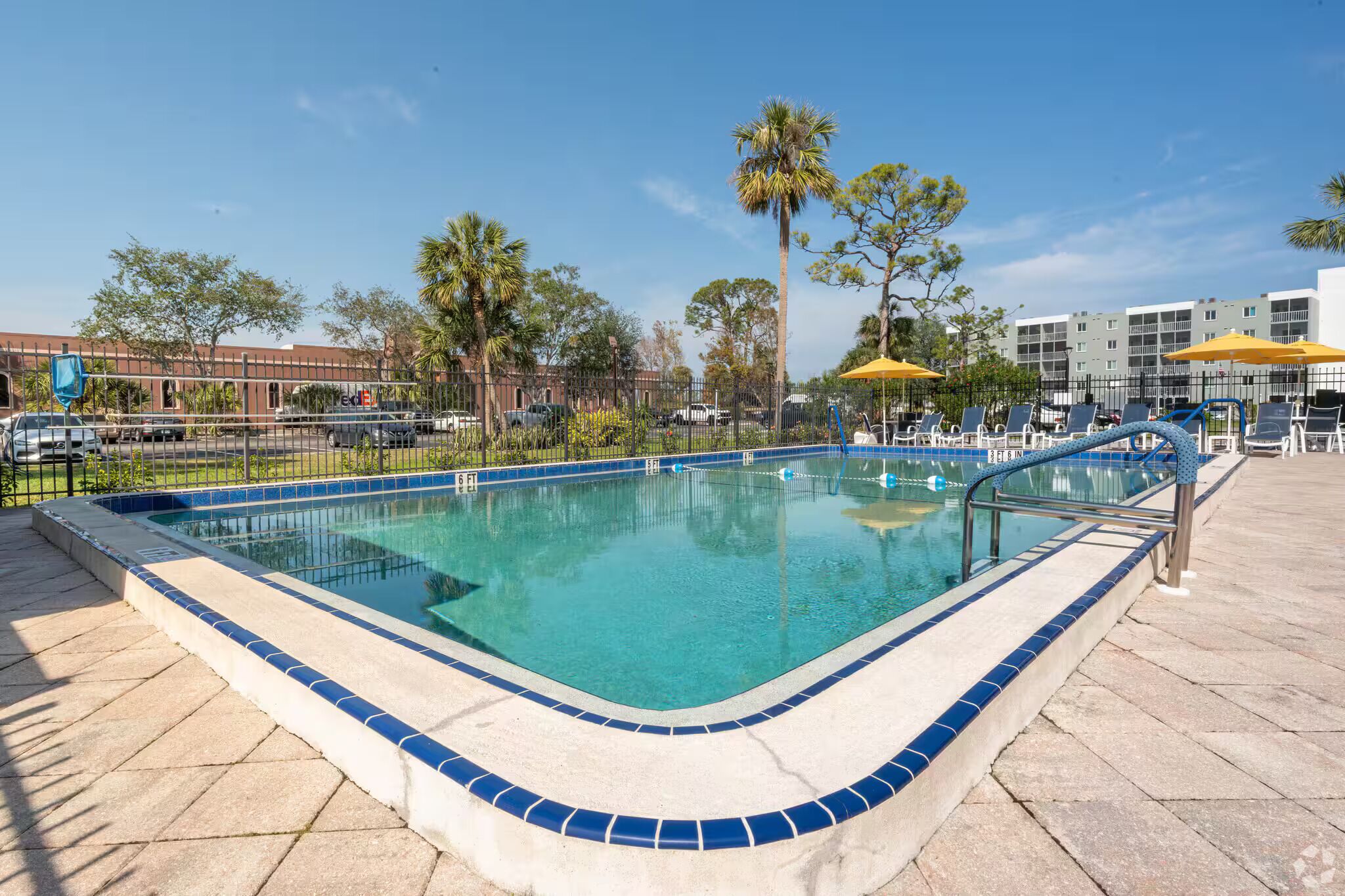 A rectangular pool with a tiled edge, surrounded by a fence, with lounge chairs and umbrellas in the background.