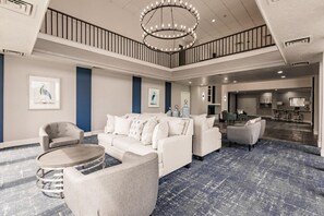 A spacious hotel lobby with a high ceiling, a chandelier, a seating area with sofas and chairs, and a visible kitchen area. - Landing Apartments Near HCA Florida Fawcett Hospital (Port Charlotte)