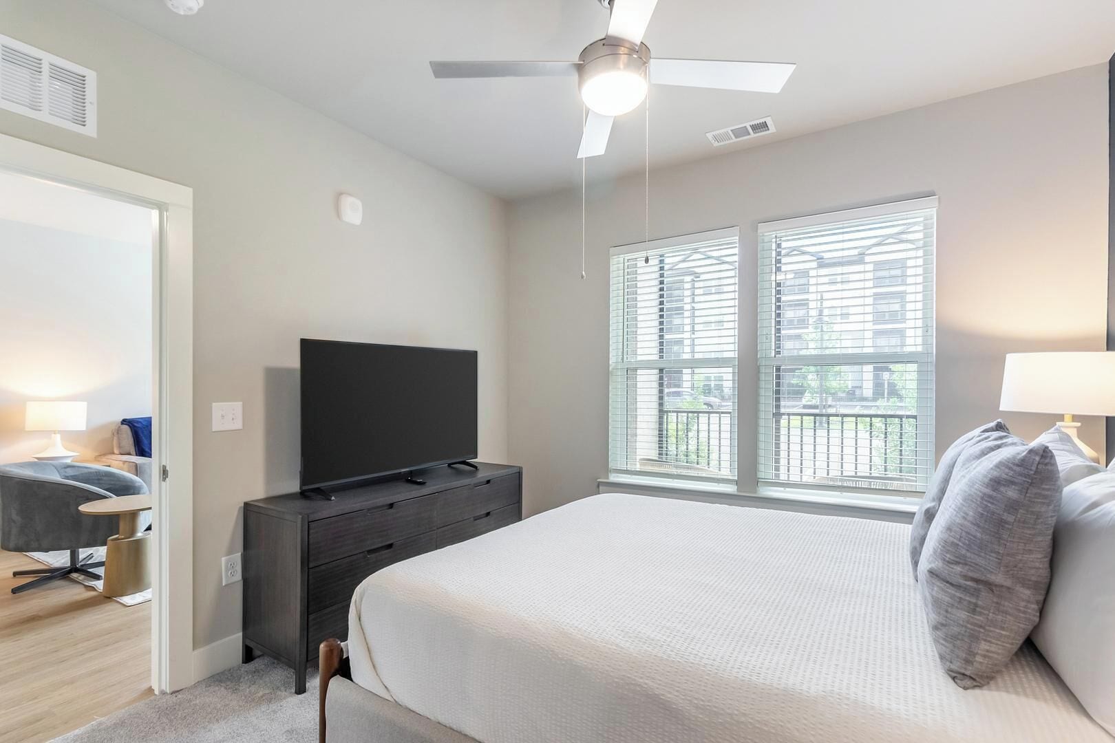 Une chambre à coucher avec un lit, un téléviseur posé sur une commode, un ventilateur de plafond et une fenêtre avec des stores.