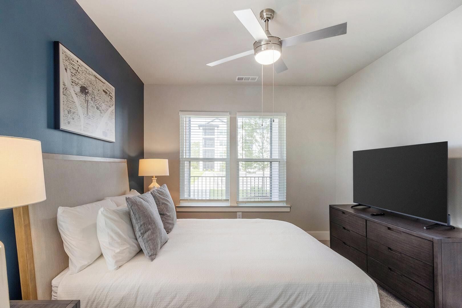 A bedroom with a bed, a ceiling fan, a TV, a dresser, and a window with blinds.